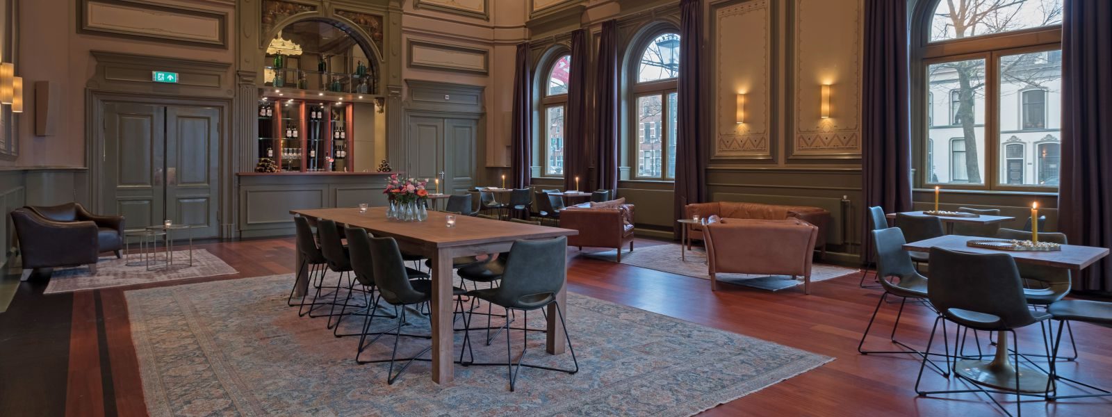 Een historische zaal met hoge boogramen, houten lambrisering, een lange tafel met stoelen en leren banken
